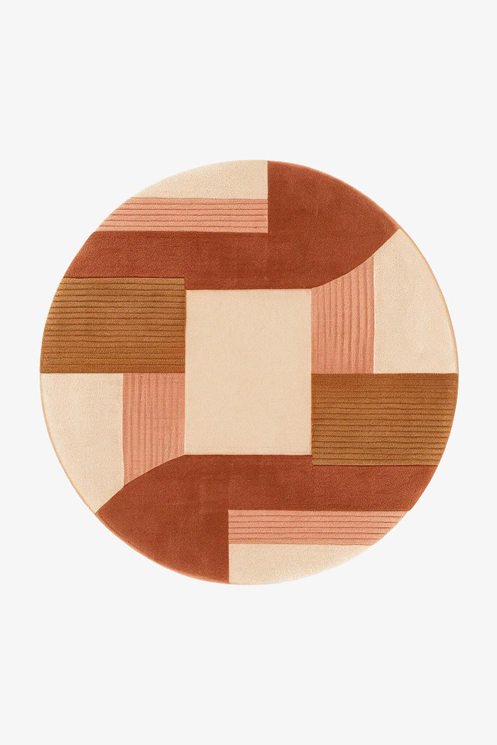 A round area rug with a geometric patchwork design in terracotta, dusty pink, beige, and brown, shown from a top-down view on a light gray background.