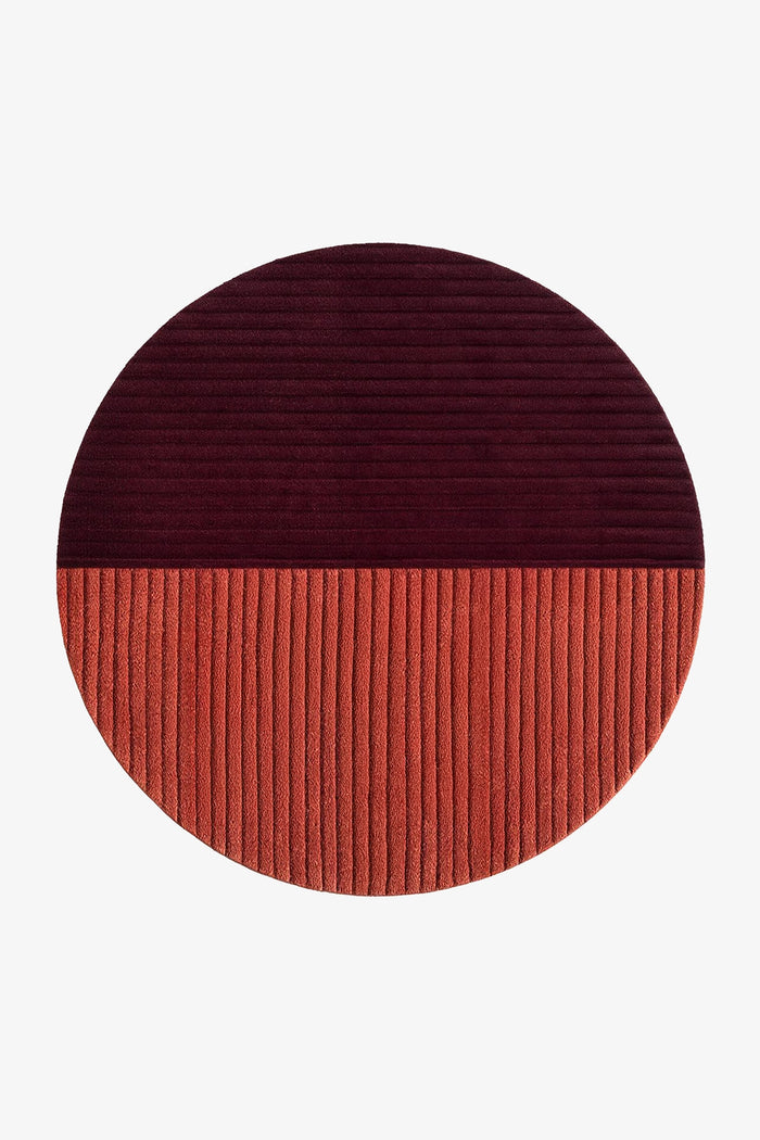 A round ribbed rug split horizontally into two colors: a dark burgundy top half and a terracotta orange bottom half, shown on a plain white background rust