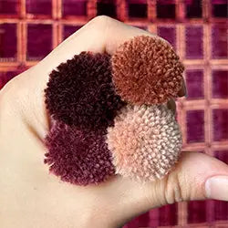 Three pom-poms in purple, brown, and pink held in a hand against a patterned rug background.