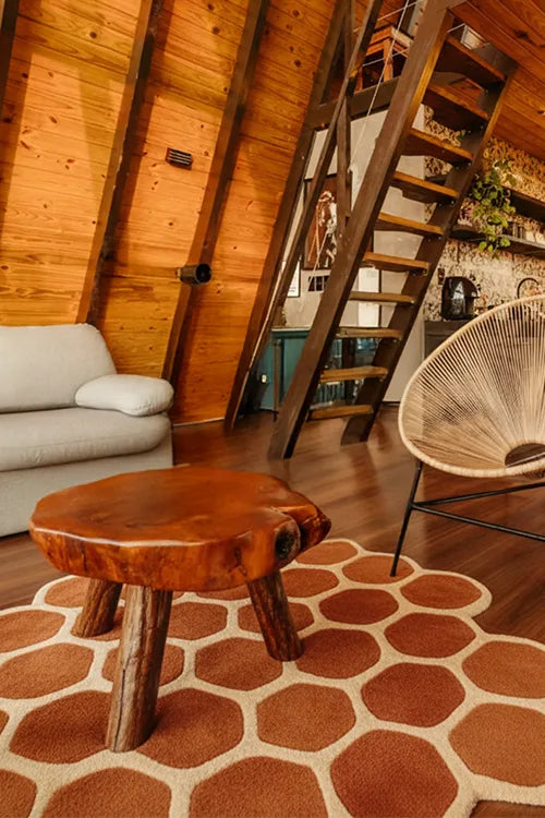 Wooden stool on a unique graphic honeycomb rug in a room with a sloped wooden ceiling, staircase, and modern furniture.