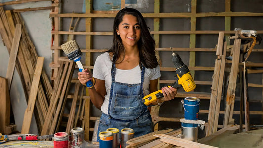 photo of A woman holding various tools, such as a drill, hammer, and paintbrush, with materials, like wood planks and paint cans, scattered around them in a workspace