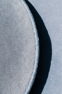 Close-up of a light baby blue circular rug with a stitched edge, shown against gray backing with a strong shadow | Cotton backing