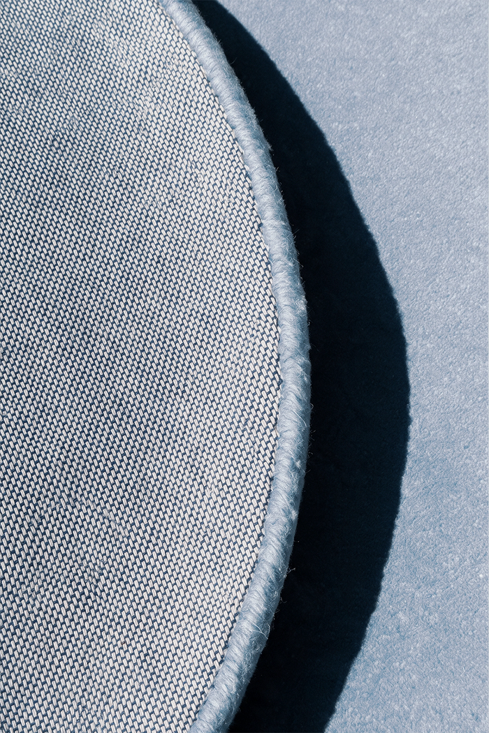 Close-up of a light baby blue circular rug with a stitched edge, shown against gray backing with a strong shadow | Cotton backing