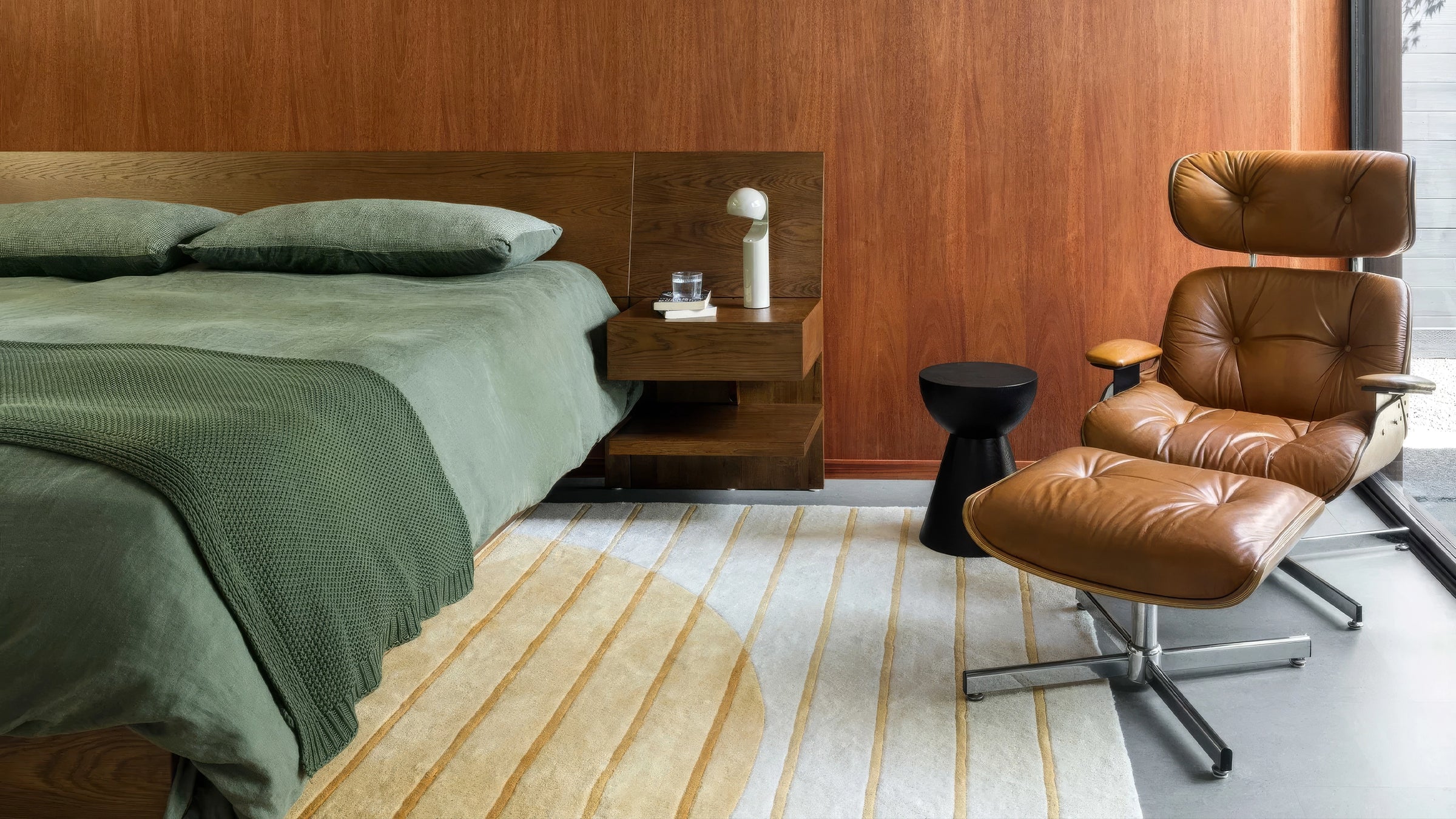 A mid-century modern brown leather tufted lounge chair with an ottoman in a styled bedroom setting with a wood-paneled wall, bed, and striped rug.
