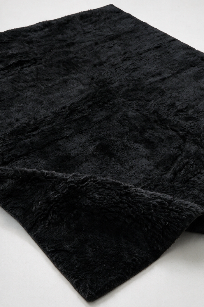 A close-up of a black shaggy wool rug, showing its soft, plush texture and folded corner on a plain white background.