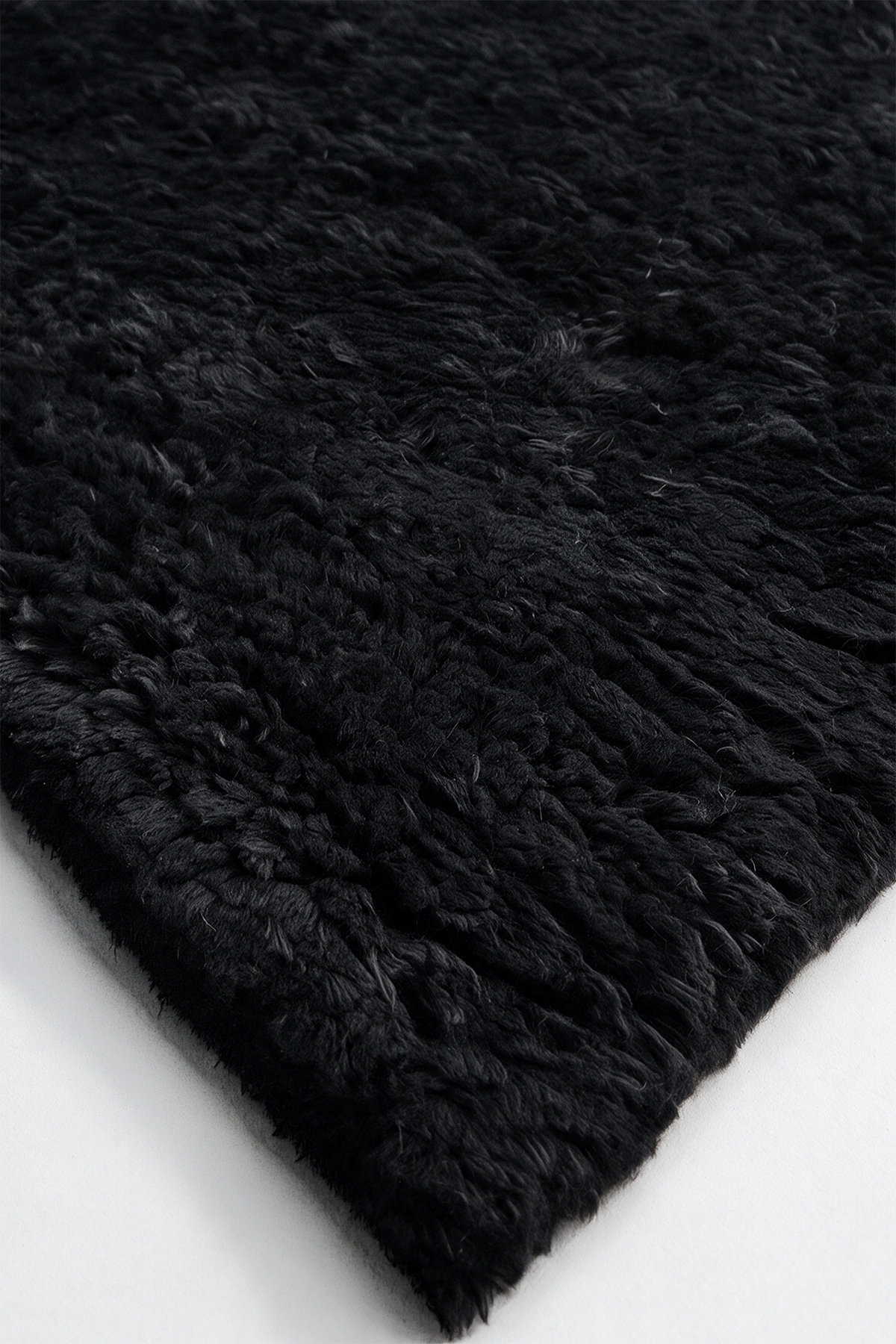 A close-up of a black shaggy wool rug showing its thick, textured pile and folded corner on a white surface.