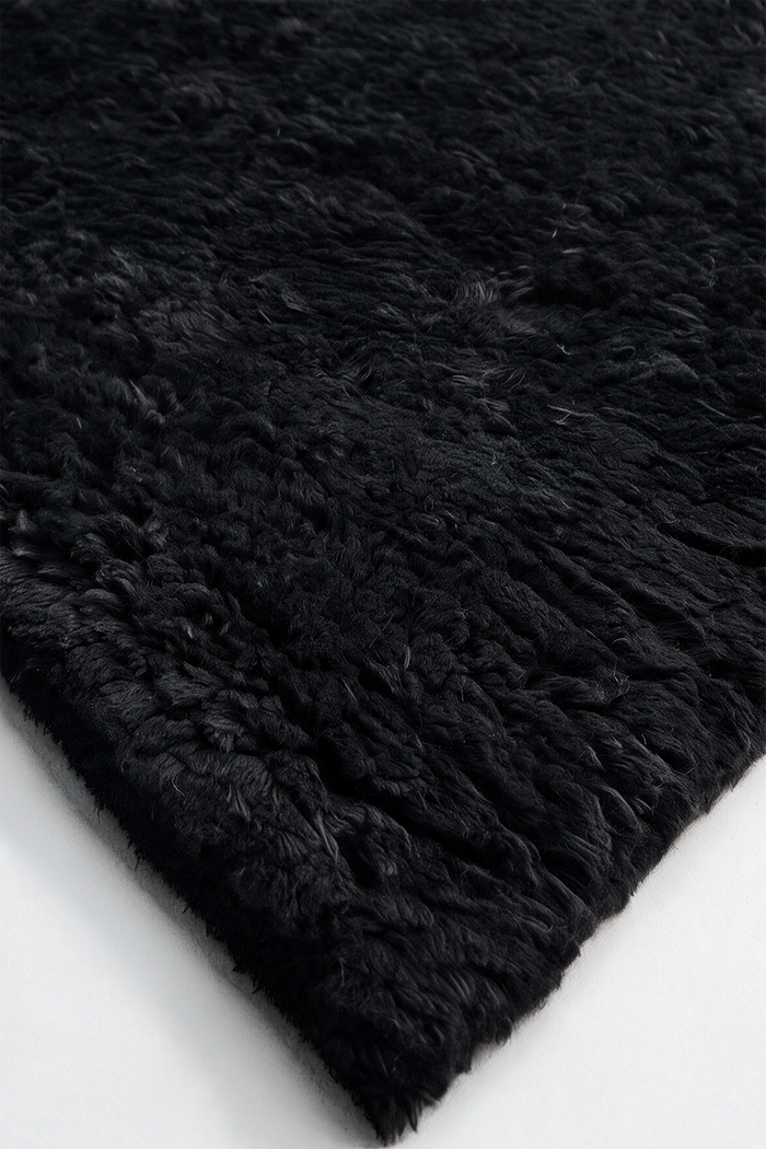 A close-up of a black shaggy wool rug showing its thick, textured pile and folded corner on a white surface.