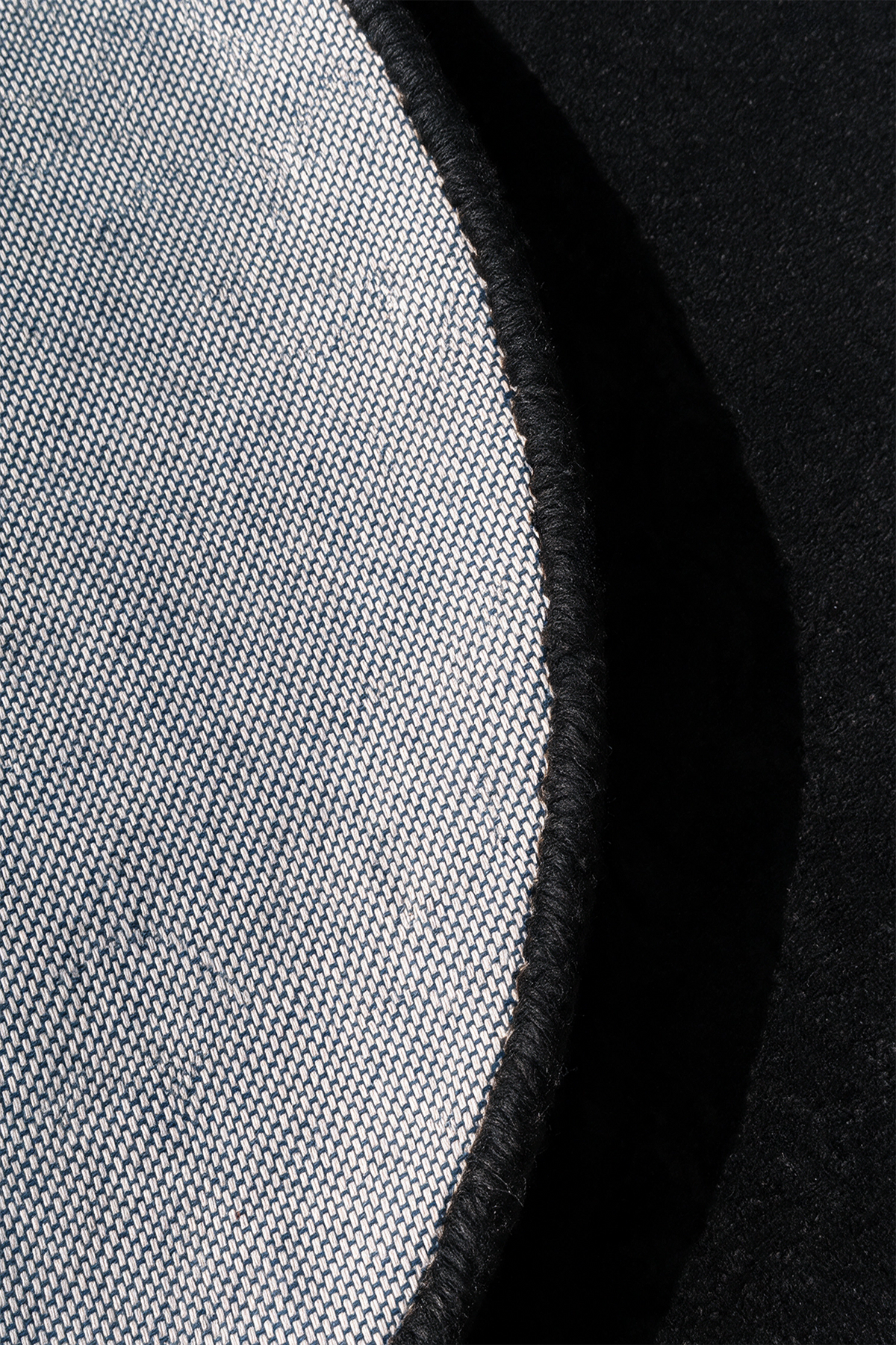 Close-up detail of a circular rug showing a light gray woven fabric texture with a black stitched edge against a black background.  | Cotton backing