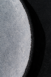 Close-up detail of a circular rug showing a light gray woven fabric texture with a black stitched edge against a black background.  | Cotton backing