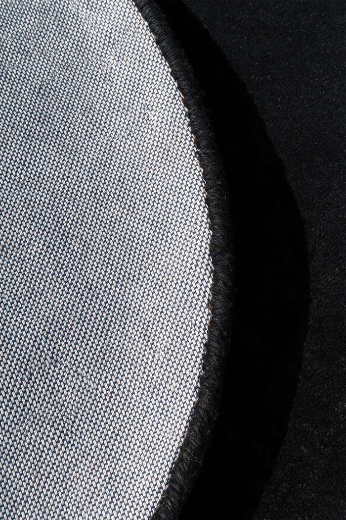 Close-up detail of a circular rug showing a light gray woven fabric texture with a black stitched edge against a black background.