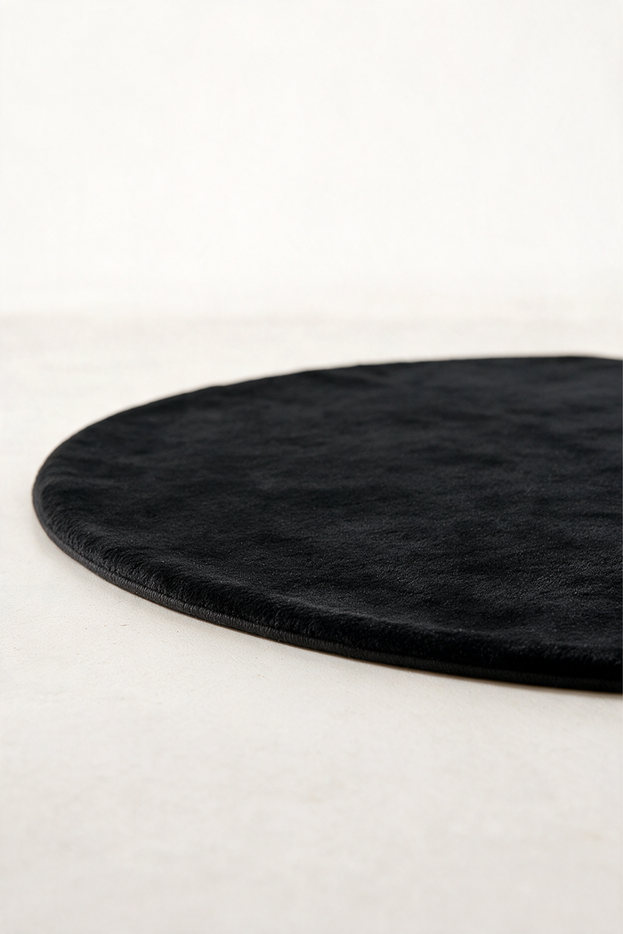A close-up of a round black rug with a soft, plush texture, cropped to show its curved edge on a light beige floor against a white wall.