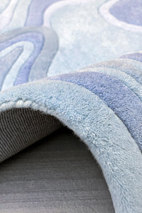 A close-up of a plush area rug with a wavy, organic pattern in shades of light blue, and gray, shown with one corner folded over to reveal the texture and underside.