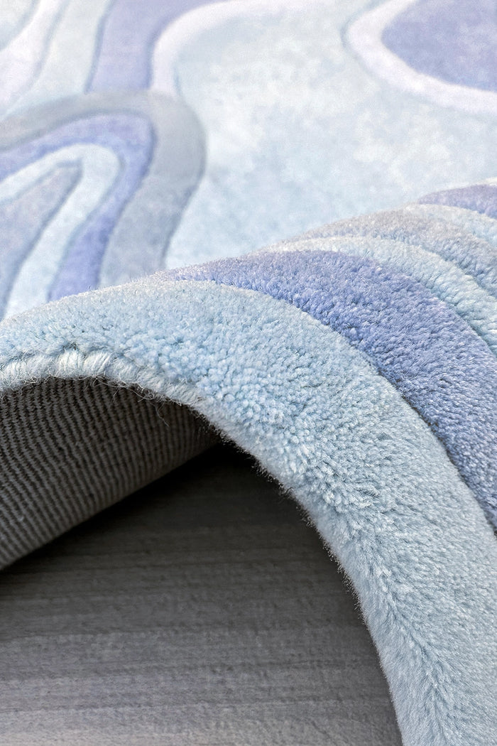 A close-up of a plush area rug with a wavy, organic pattern in shades of light blue, and gray, shown with one corner folded over to reveal the texture and underside.