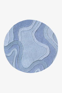 A round area rug featuring an abstract, layered topographic pattern in various shades of blue and light gray, shown against a plain light gray background.