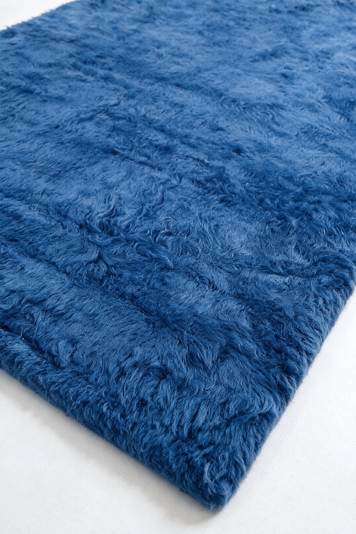 A close-up of a plush, shaggy blue rug corner showing its thick, curly texture against a clean white background.