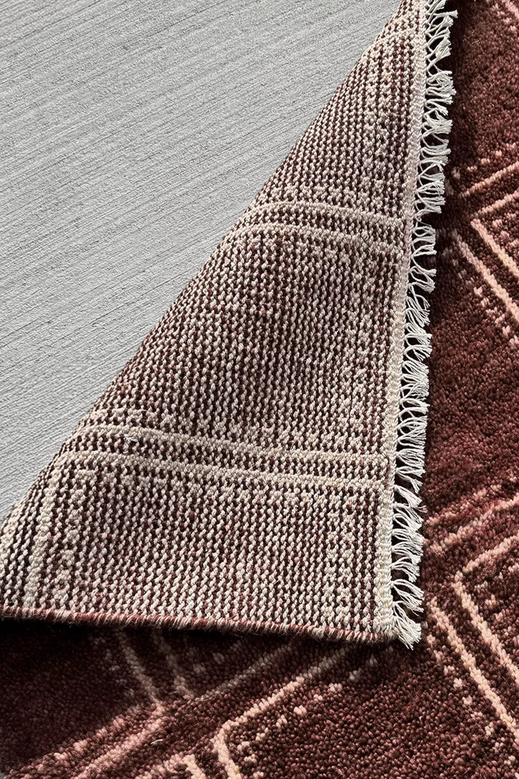 Close-up of a woven rug corner showing a textured pattern in taupe and brown with fringed edges, placed on a light gray floor over a darker brown rug surface.