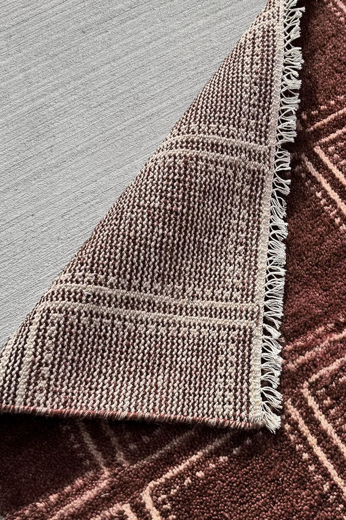 Close-up of a woven rug corner showing a textured pattern in taupe and brown with fringed edges, placed on a light gray floor over a darker brown rug surface.