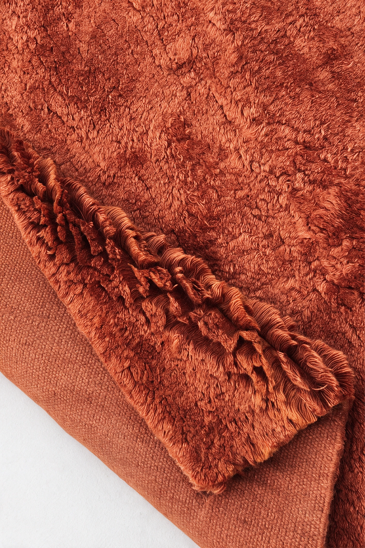 A close-up of a rust-orange shaggy rug with a contrasting textured woven border, folded over to show its dual-texture surface burnt orange