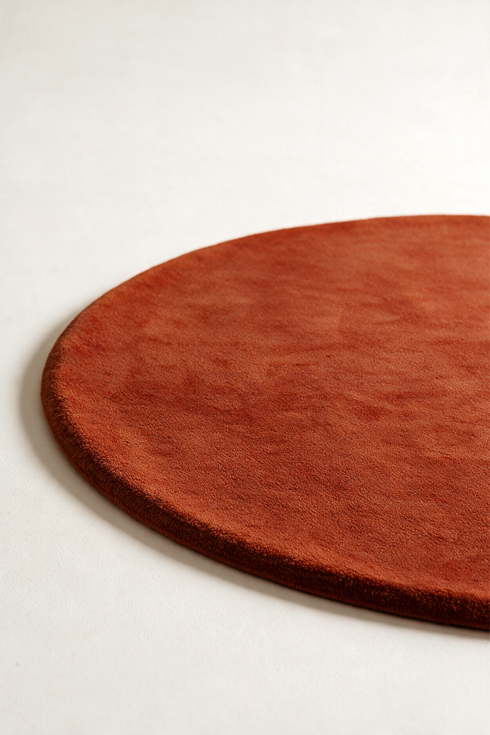 A close-up of a round rust burnt orange textured rug on a light gray background.