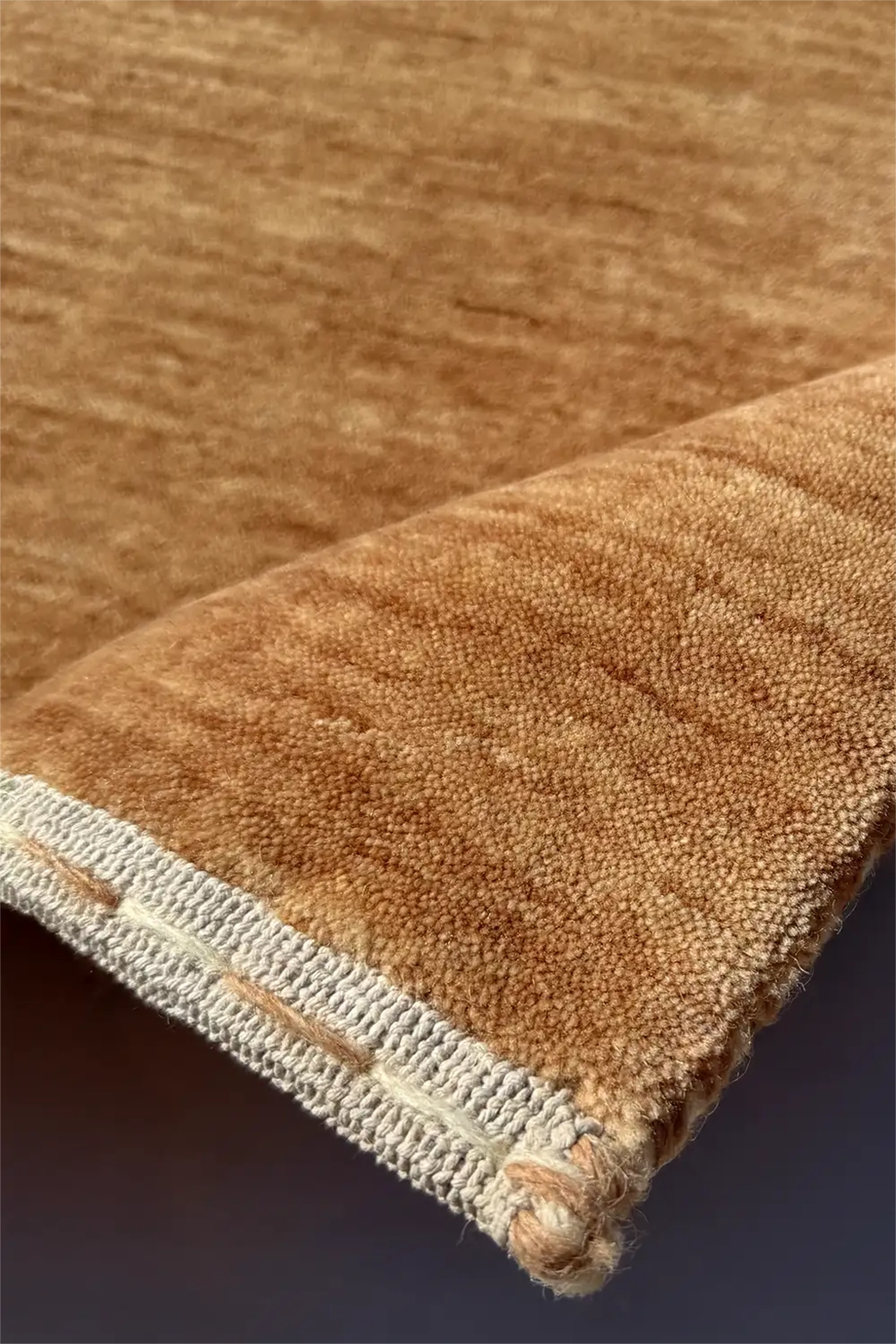 A close-up of a brown textured rug with a folded corner showing a cream-colored woven edge camel texture