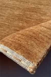 A close-up of a brown textured rug with a folded corner showing a cream-colored woven edge camel texture
