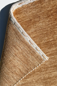 Close-up of a woven runner rug corner showing a warm tan and brown textured surface with a cream stitched border camel