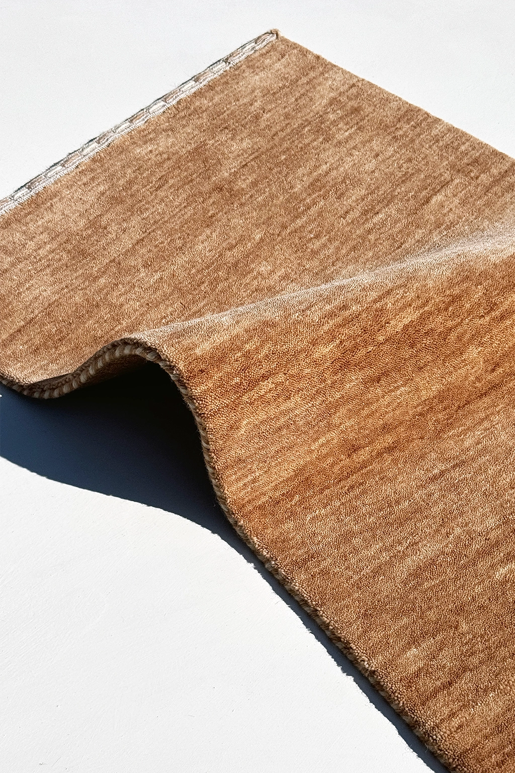 A close-up of a textured runner rug with a visible woven edge, folded over to show its thickness and backing, set against a plain white background camel texture
