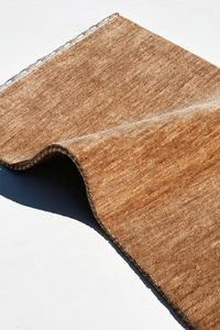 A close-up of a textured runner rug with a visible woven edge, folded over to show its thickness and backing, set against a plain white background camel texture