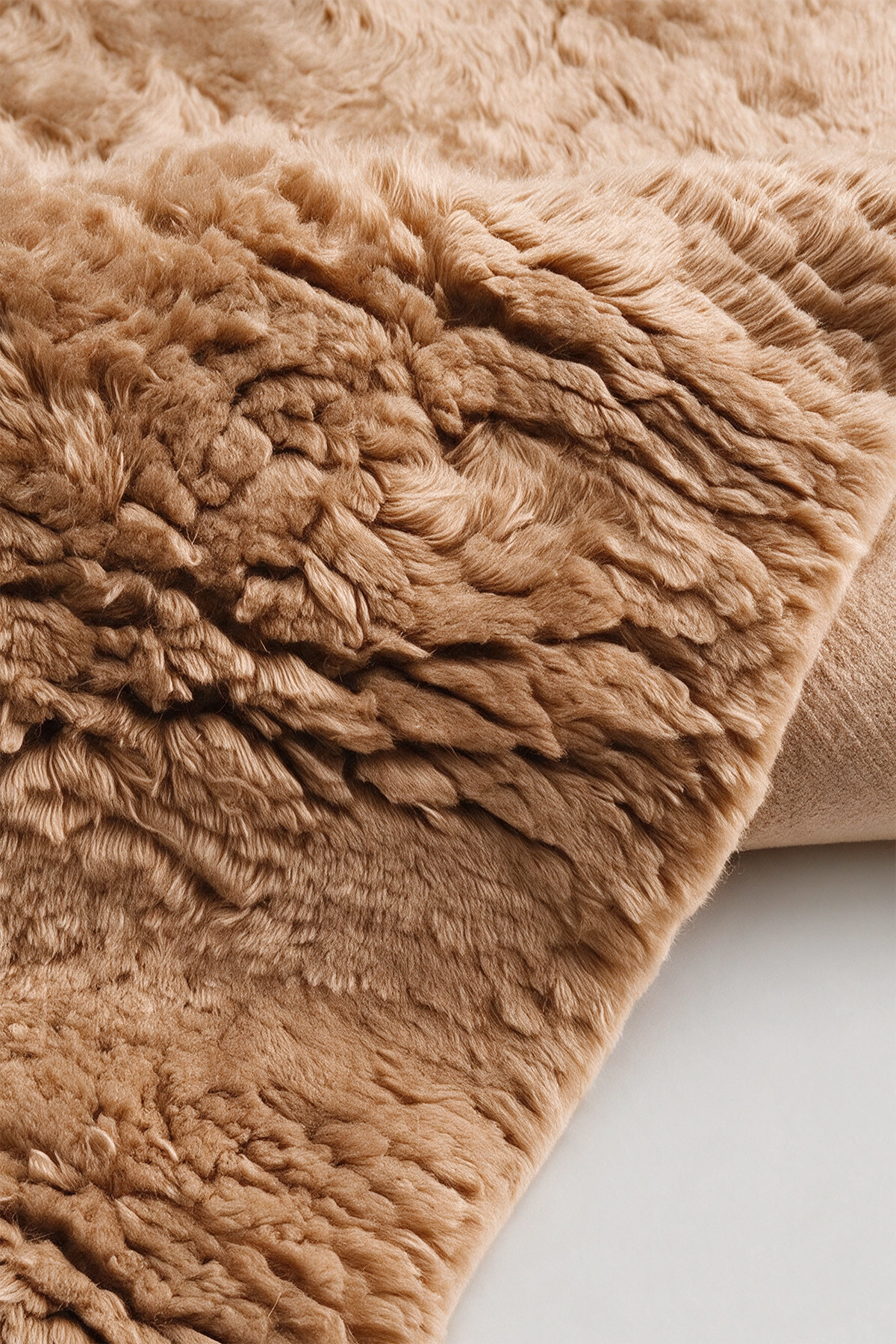 A close-up of a plush, shaggy wool rug in a warm tan or camel color, folded over to show its thick texture against a light neutral background.