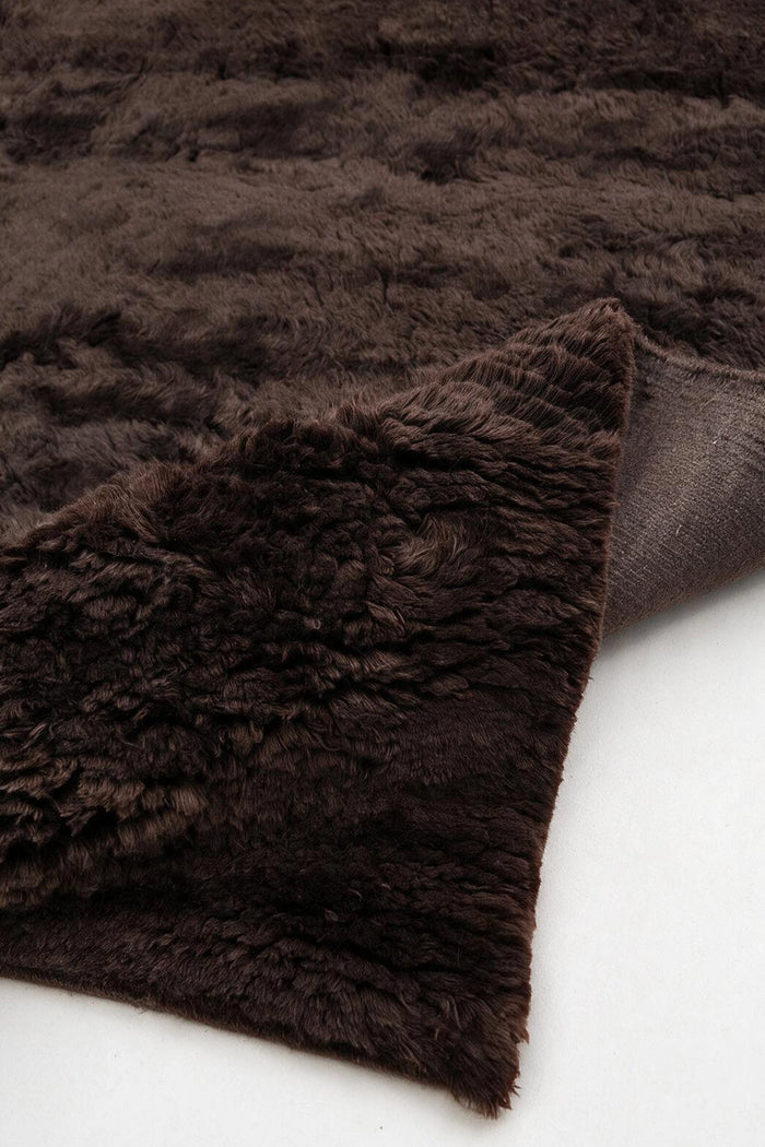 A close-up of a dark brown shaggy wool  rug, showing its thick textured pile and folded corner revealing the brown knotted backing, set against a plain white background