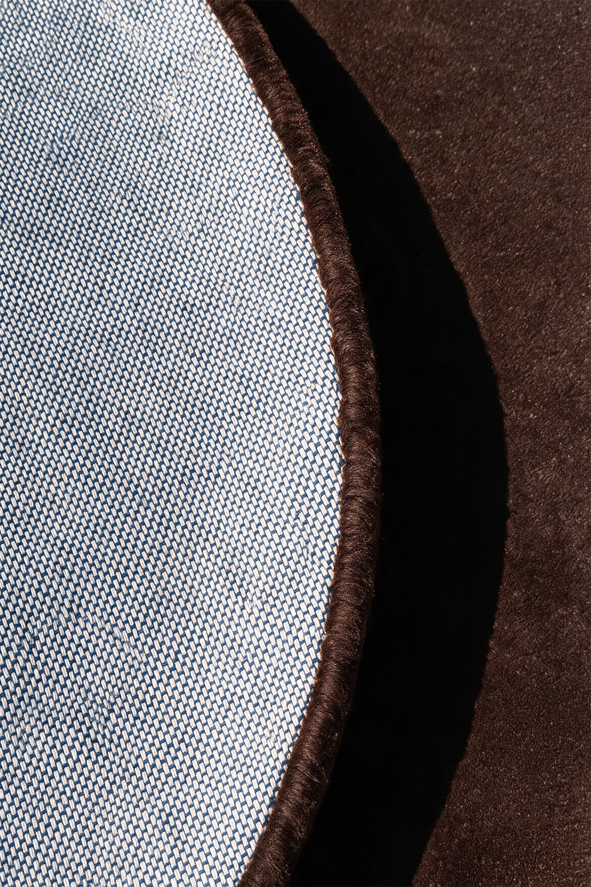 Close-up detail of a round rug showing a light blue-gray woven fabric edge with a dark brown tufted border, set against a dark chocolate brown textured surface | Cotton backing