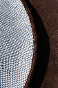 Close-up detail of a round rug showing a light blue-gray woven fabric edge with a dark brown tufted border, set against a dark chocolate brown textured surface | Cotton backing