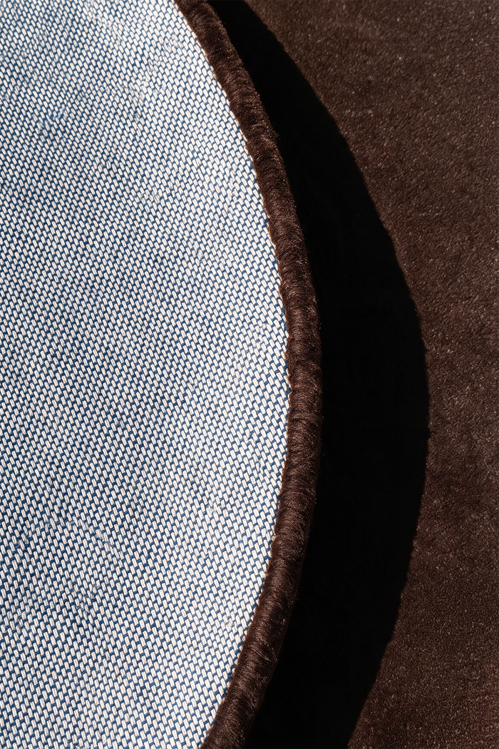 Close-up detail of a round rug showing a light blue-gray woven fabric edge with a dark brown tufted border, set against a dark chocolate brown textured surface | Cotton backing