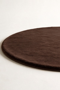 A close-up of a dark chocolate brown round rug with a soft, plush texture, shown on a clean off-white background.
