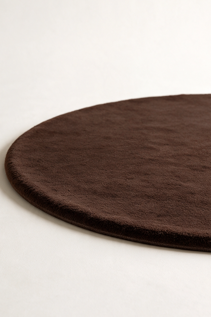 A close-up of a dark chocolate brown round rug with a soft, plush texture, shown on a clean off-white background.