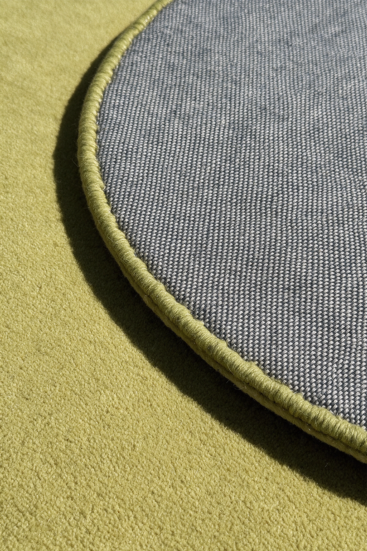 Close-up of a round rug showing two colorways: a textured olive green top surface and a gray woven underside with a matching citron green stitched edge.