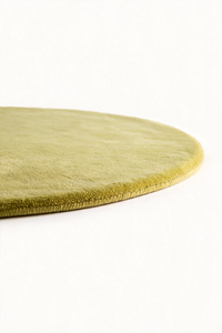 A close-up of an oval area rug in a muted citron color, showing its plush texture and rolled edge against a plain white background.
