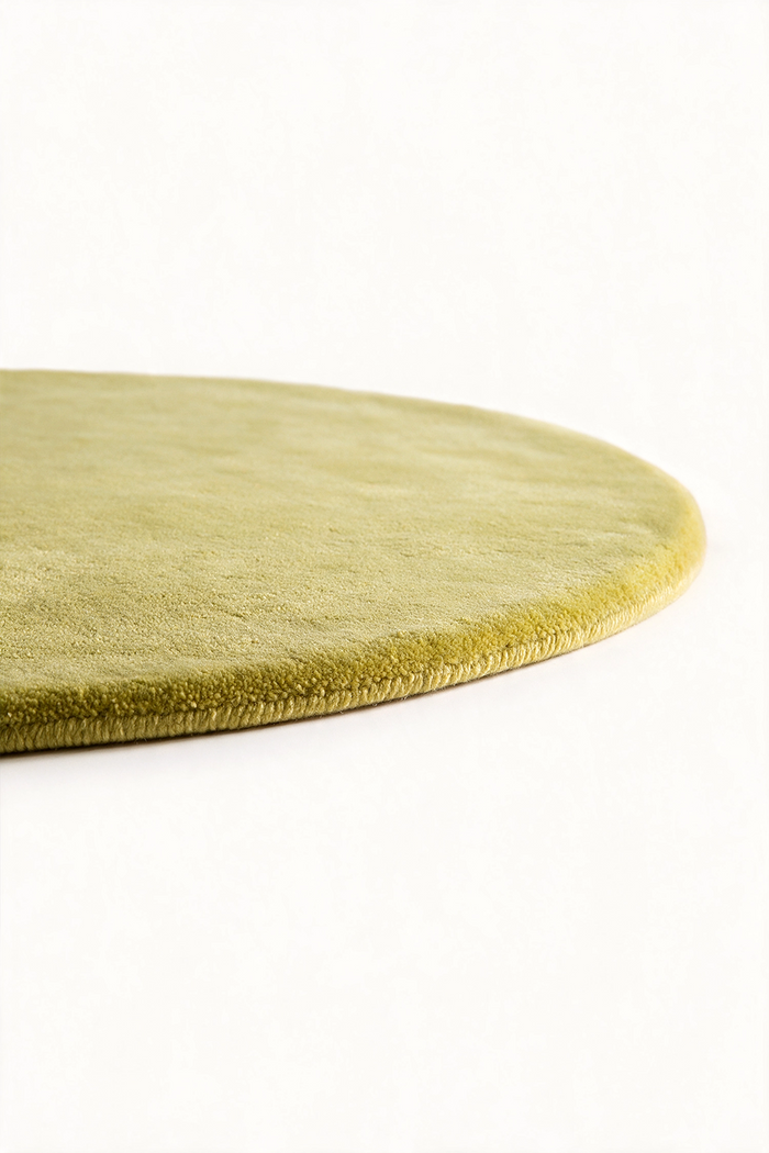 A close-up of an oval area rug in a muted citron color, showing its plush texture and rolled edge against a plain white background.