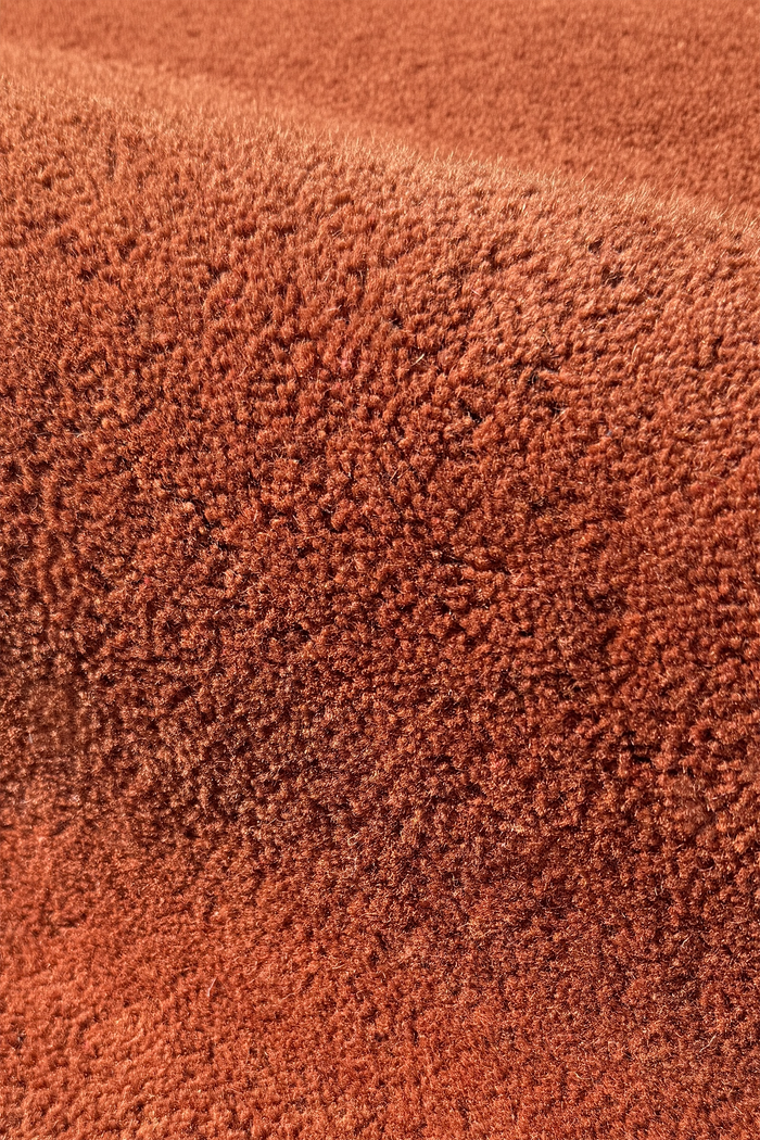 A close-up of a textured terracotta or rust-colored wool rug, showing the dense pile and soft folds brown