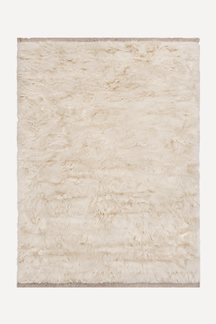 A rectangular shaggy wool rug in cream beige with fringed edges, shown flat against a light off-white background.