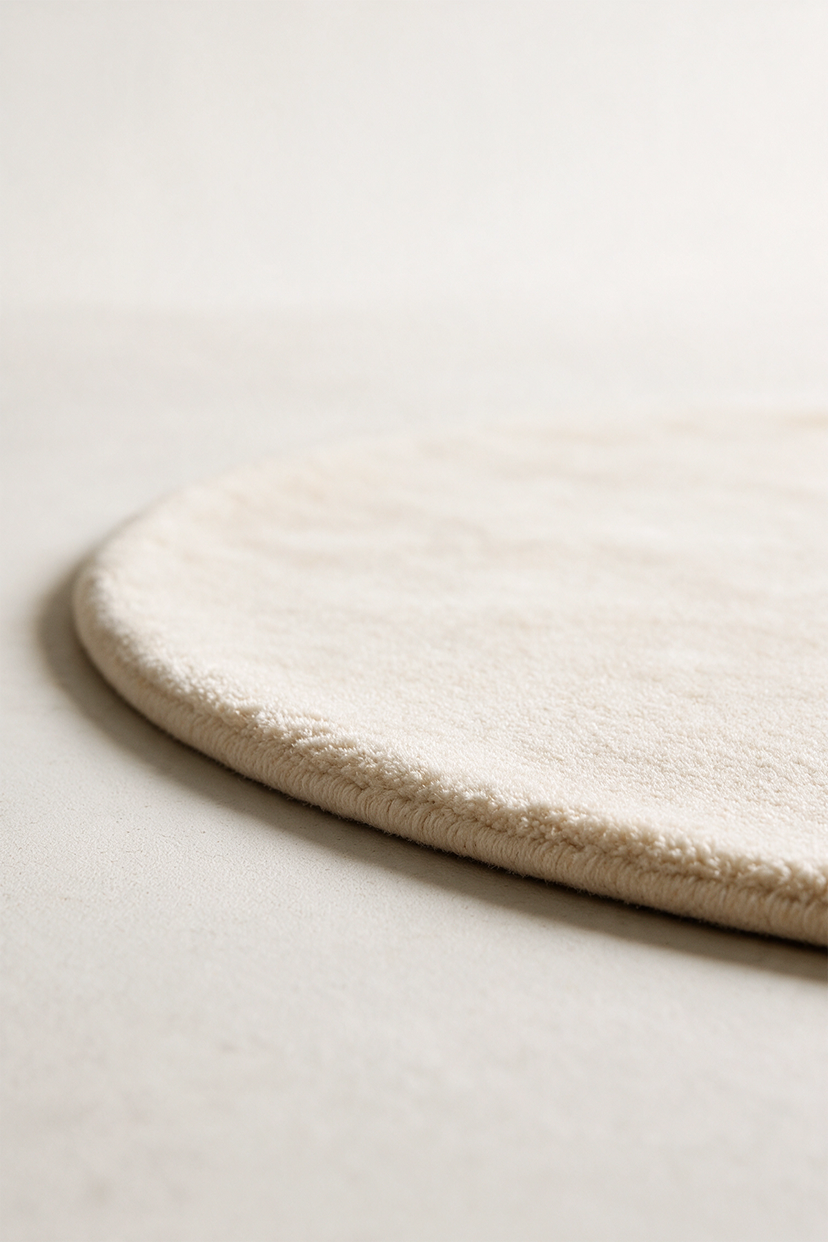 A close-up of a round, plush cream-colored rug with a thick tufted edge, resting on a neutral light beige surface.