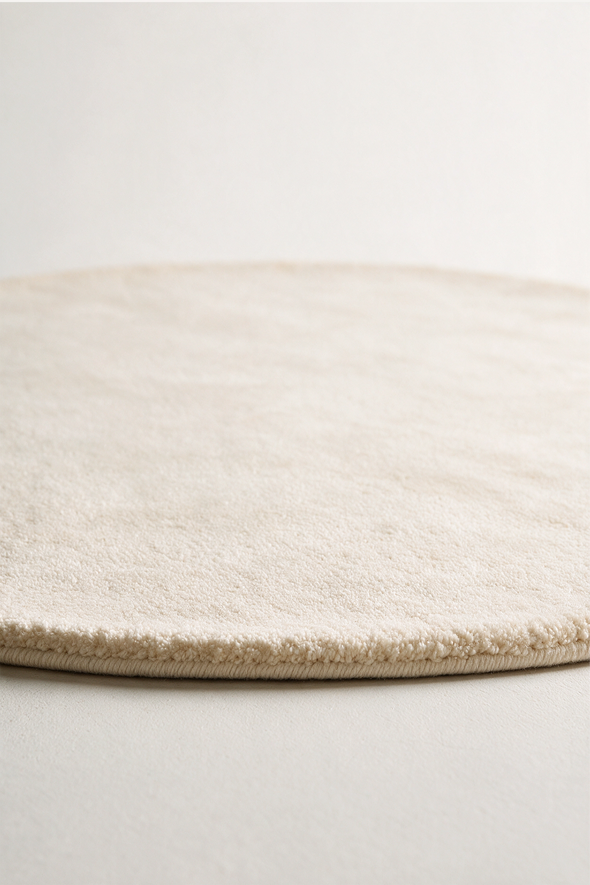 A close-up of a round, low-pile area rug in a warm cream or off-white color, showing the soft texture and short looped pile on a neutral light gray background.
