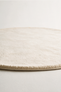 A close-up of a round, low-pile area rug in a warm cream or off-white color, showing the soft texture and short looped pile on a neutral light gray background.