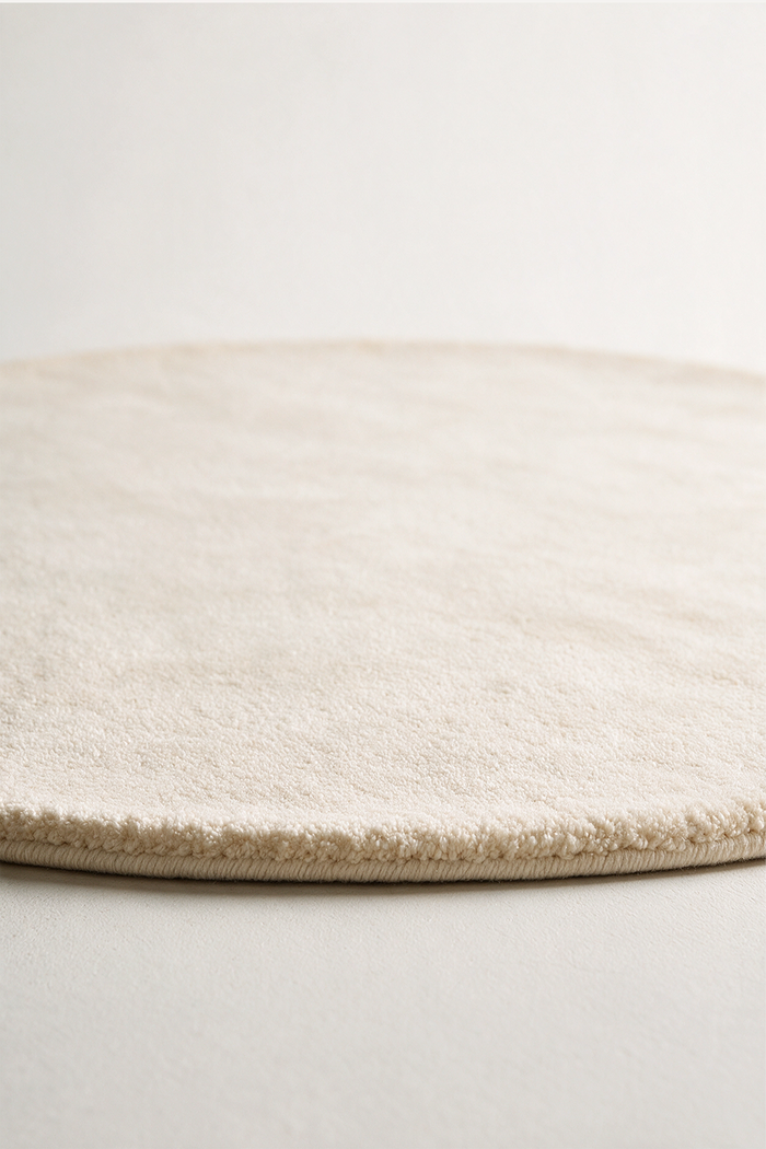 A close-up of a round, low-pile area rug in a warm cream or off-white color, showing the soft texture and short looped pile on a neutral light gray background.