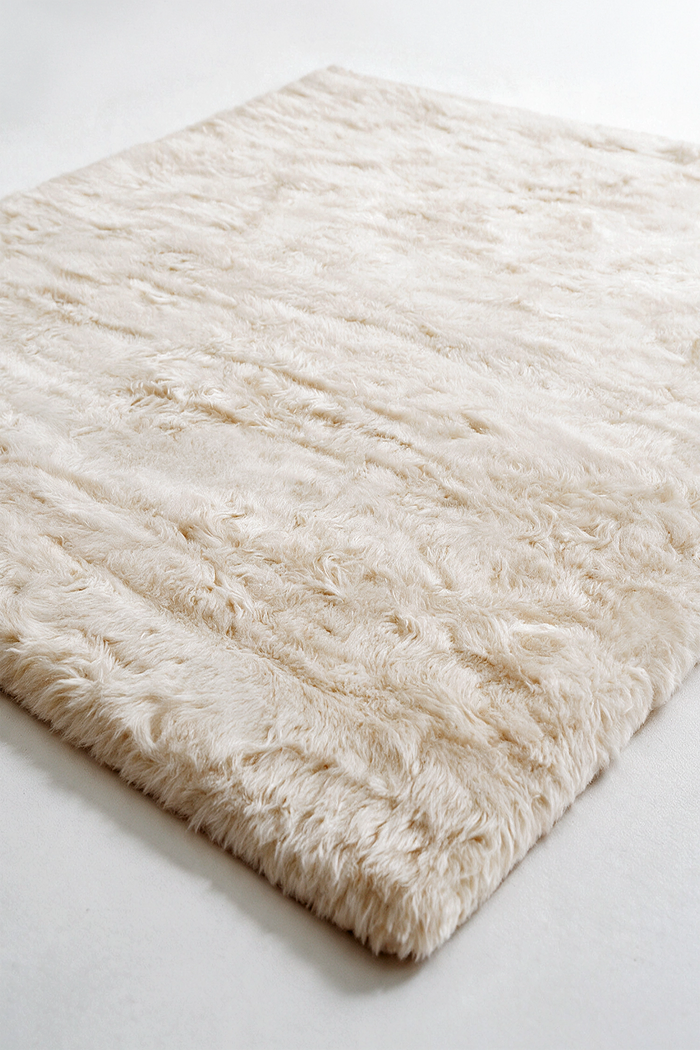 A close-up of a fluffy, shaggy area rug in a warm cream or off-white color, shown from an elevated angle on a light gray surface.