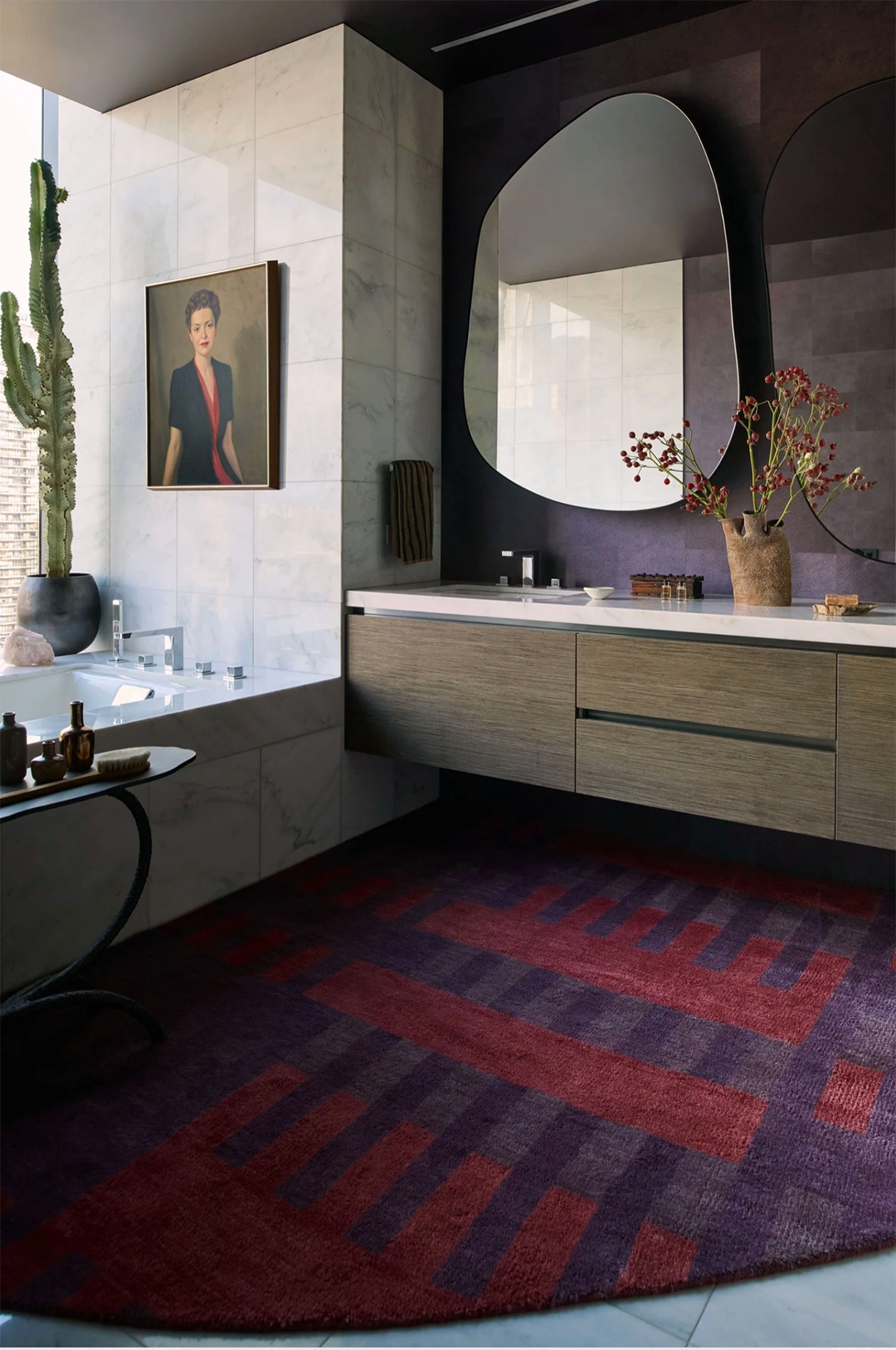 A modern bathroom interior featuring a large round area rug with a red, purple, and dark blue geometric pattern in the foreground. The room includes a double vanity with light wood cabinets, a white countertop, a large organic-shaped mirror, and a bathtub in the background.