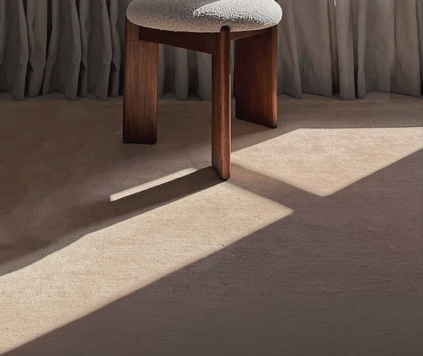 A modern wooden stool with a textured light gray upholstered seat, shown in a styled interior setting with draped curtains and sunlight casting shadows on a beige carpet.
