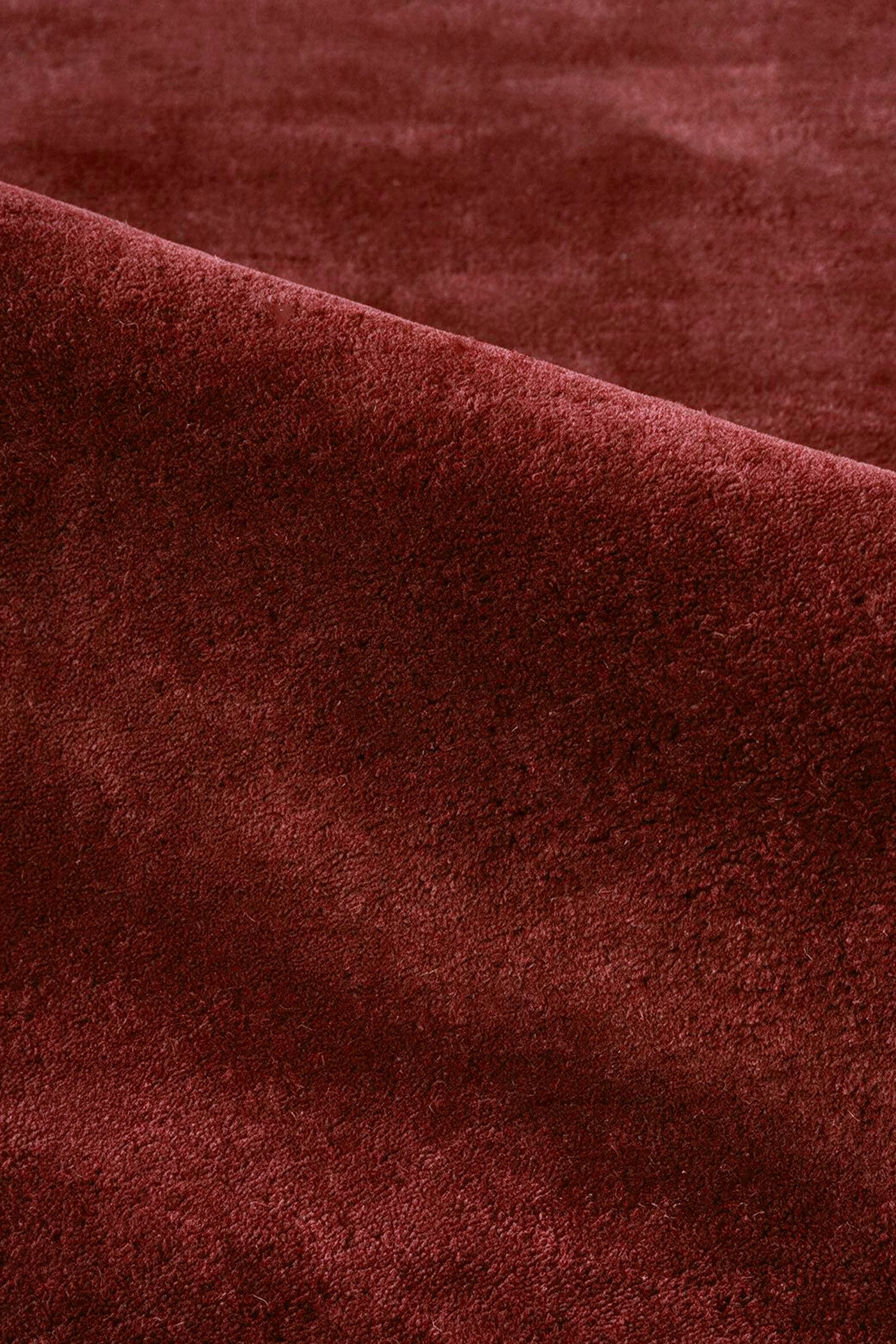 A close-up of a plush, deep burgundy rust wool rug with soft folds and a rich texture.