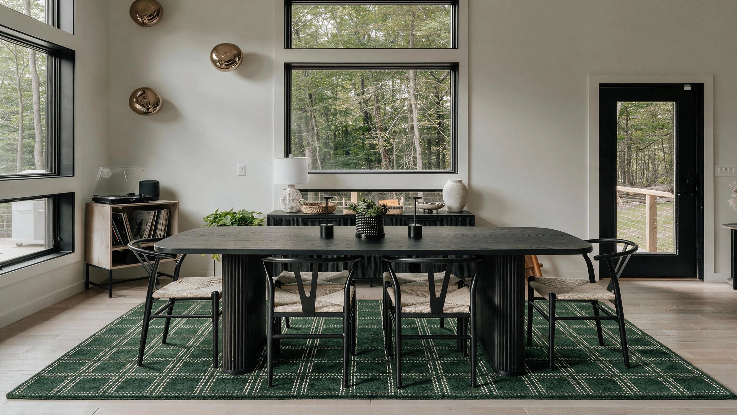 A modern dining room setup featuring a dark rectangular dining table with six chairs, placed on a green patterned area rug. The room has high windows looking out onto a forest, light-colored walls, and decorative gold wall hangings.