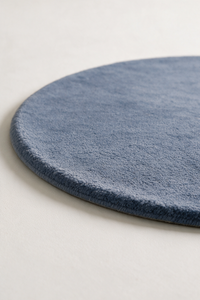 A close-up of a round dusty blue rug with a tufted pile, shown on a light neutral background.
